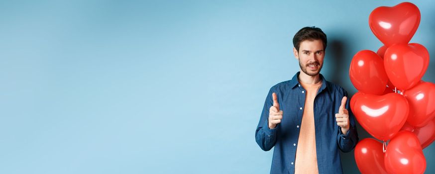 Happy valentines day. Confident man pointing fingers at camera and smiling, standing with balloons on blue background.