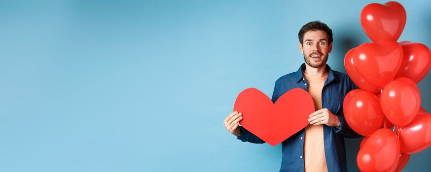 Valentines day concept. Man falling in love, looking startled at girlfriend, showing big red heart and standing near balloon over blue background.