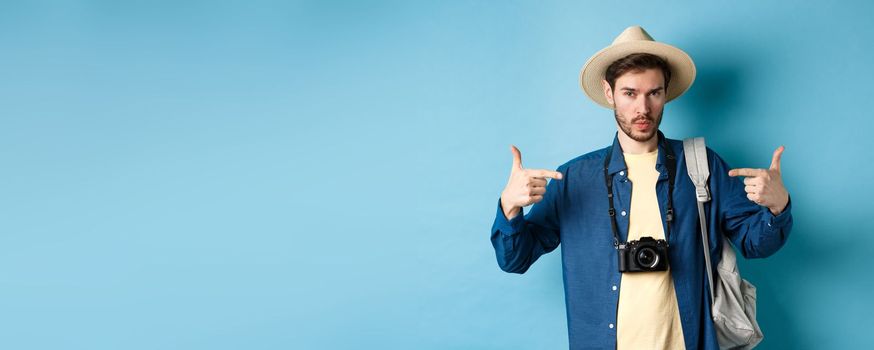 Confident and ready young man pointing at himself, self-promoting or show-off, going on summer vacation, wearing straw hat, holding backpack with camera, blue background.