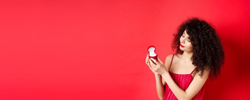 Romantic curly-haired woman in red dress, looking happy at engagement ring, become a bride, standing on studio background. Copy space