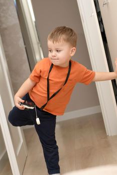 People. Handsome funny and emotional caucasian blond little boy in an orange t-shirt with a children's white camera takes pictures in the home interior. Close-up.