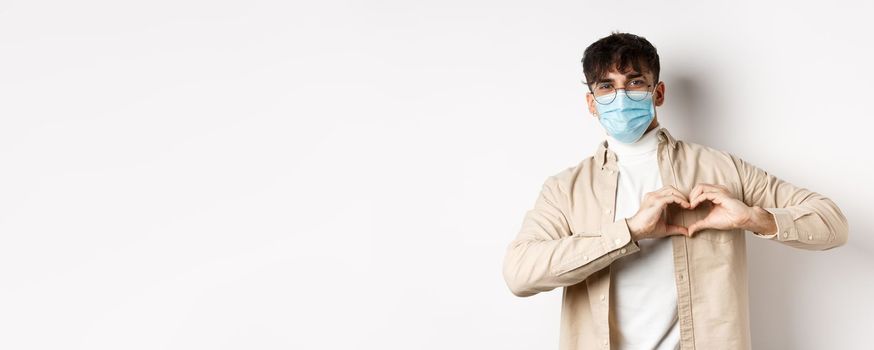 Health, covid and quarantine concept. Romantic young man in sterile medical mask showing heart gesture on chest, say I love you, standing on white background.