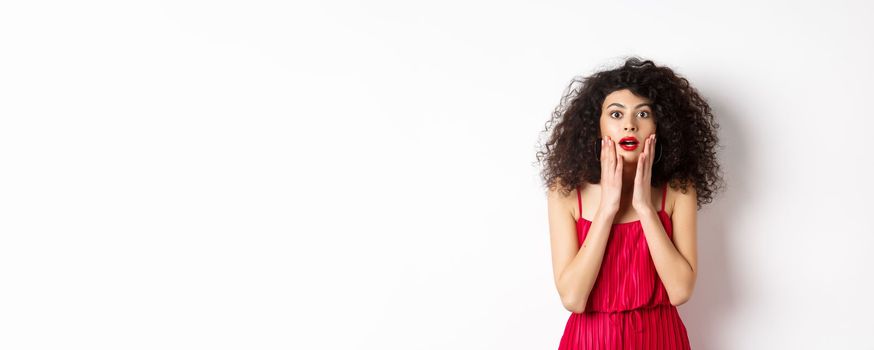 Excited curly-haired woman touch face with disbelief, looking amazed at camera, staring at promo, standing in red ress on white background.