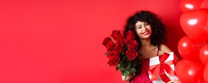 Romantic woman receive bouquet of flowers and gift on holiday, standing near heart balloons and red background, smiling grateful.