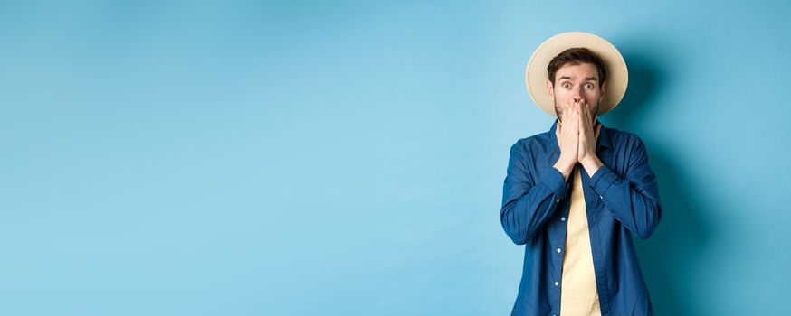 Shocked and alarmed tourist gasping, covering mouth with hands and looking startled at camera, standing on blue background.