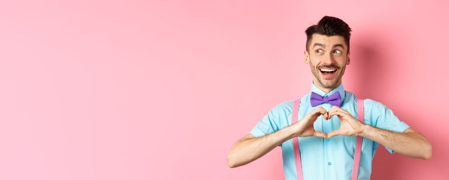Happy Valentines day. Cheerful male model enjoying romantic date, showing heart sign, say I love you and looking left with happy smile, standing over pink background.