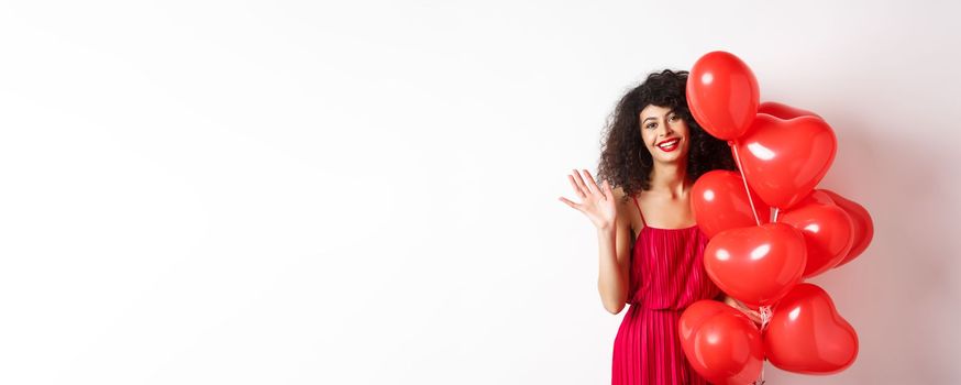 Beautiful lady with curly hair, standing near valentines holiday balloons and waving hand, saying hello, standing against white background.