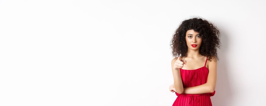 Confident young woman in red dress and makeup, pointing at camera, choosing you, inviting to event, standing over white background.