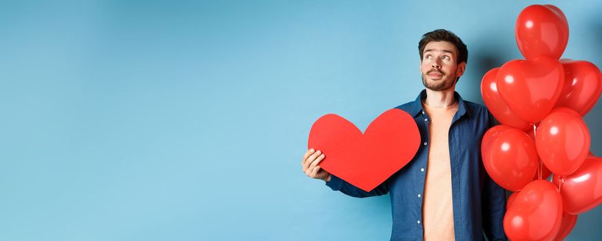 Valentines day and love concept. Man dreaming of soulmate, holding big red heart cutout and balloons, standing over blue background.