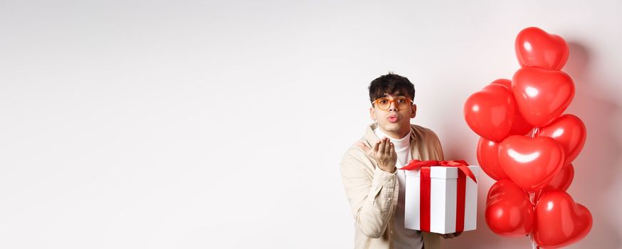 Valentines day and romance concept. Romantic modern man holding special gift for lover and sending air kiss at camera, standing near hearts balloons, white background.