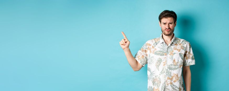 Disappointed tourist cringe and frown, pointing finger aside at empty space, complaining on bad thing, standing in hawaiian shirt on blue background.