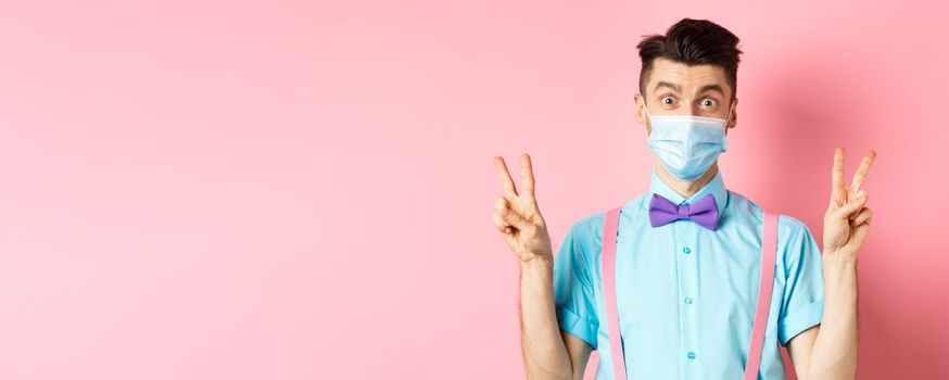 Coronavirus, healthcare and quarantine concept. Funny guy in bow-tie and face mask showing peace, victory signs and looking happy at camera, standing on pink background.