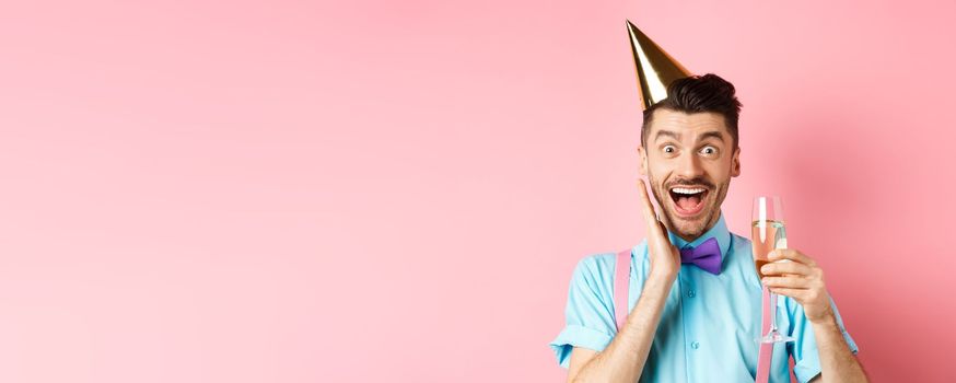 Holidays and celebration concept. Funny young man in birthday hat celebrating, screaming from joy and surprise, raising glass of champagne and smiling, standing on pink background.