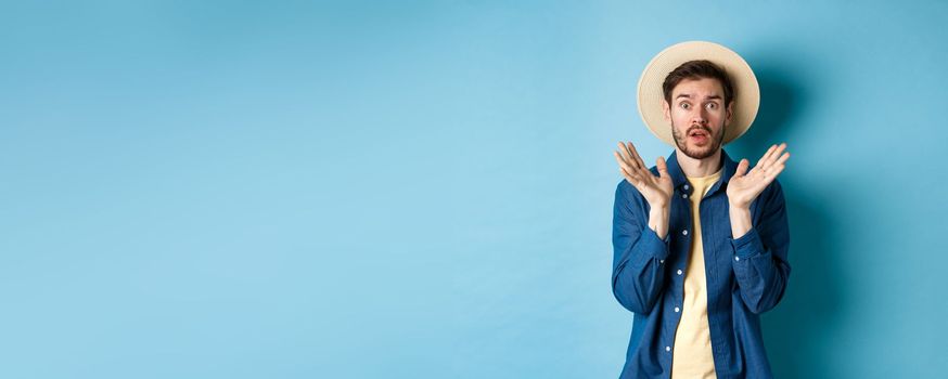 Image of shocked tourist in straw hat panicking, raising hands up and stare startled at camera, standing on blue background.