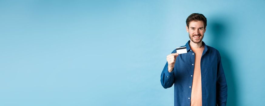 Handsome smiling guy showing plastic credit card and looking satisfied, standing on blue background.