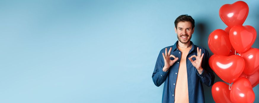 Happy valentines day. Cheerful boyfriend showing okay gestures and praise something good, standing with red hearts balloons for lover, blue background.