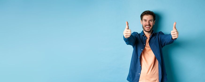 Handsome young man smiling and showing thumbs up in approval, say yes with happy face, standing on blue background.