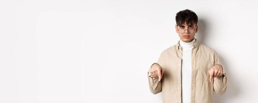 Portrait of handsome hipster guy in glasses showing an ad, pointing fingers down at logo, standing on white background.