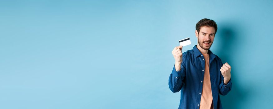 Satisfied young man showing plastic credit card and say yes, cheering of joy, standing on blue background.