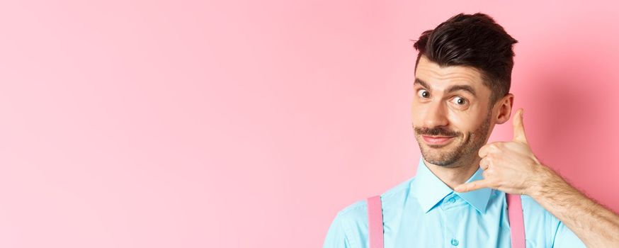 Close-up of handsome young guy with moustache, showing phone gesture, asking to call him, standing on pink background.
