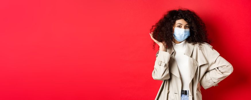 Covid-19, pandemic and quarantine concept. Stylish coquettish woman in medical mask and trench coat, fixing her curly hairstyle and smiling, red background.