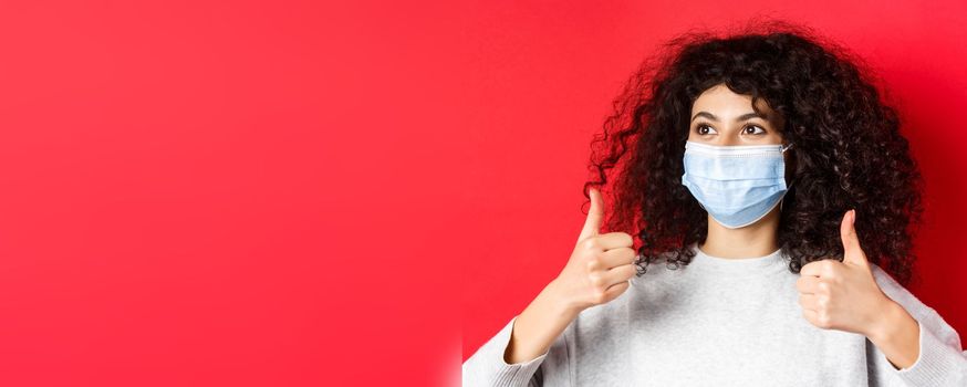 Covid-19 and pandemic concept. Close-up of young caucasian woman with curly hair, wearing medical mask, showing thumbs up and looking aside at empty space, red background.