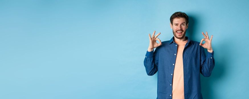 Happy smiling man winking and showing alright OK sign, say yes, approve and agree to something good, standing on blue background.