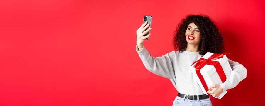 Happy young woman taking selfie with her valentines day gift, holding present and photographing on smartphone, posing on red background.