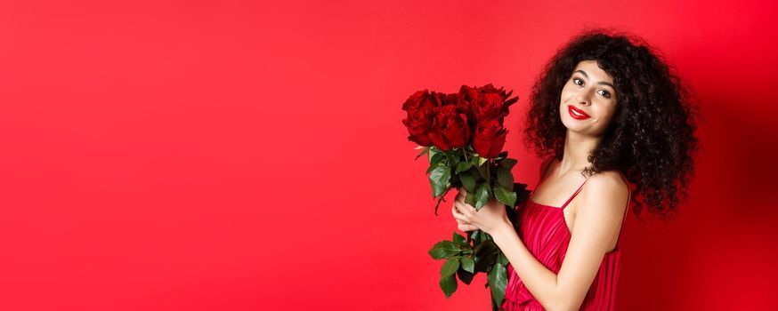 Happy beautiful woman in dress, holding flowers and smiling romantic, standing against red background.