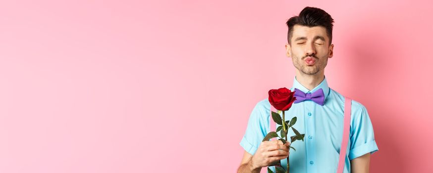 Cute and funny man waiting for kiss from lover on Valentines day, holding beautiful red rose for girlfriend, standing over pink background.