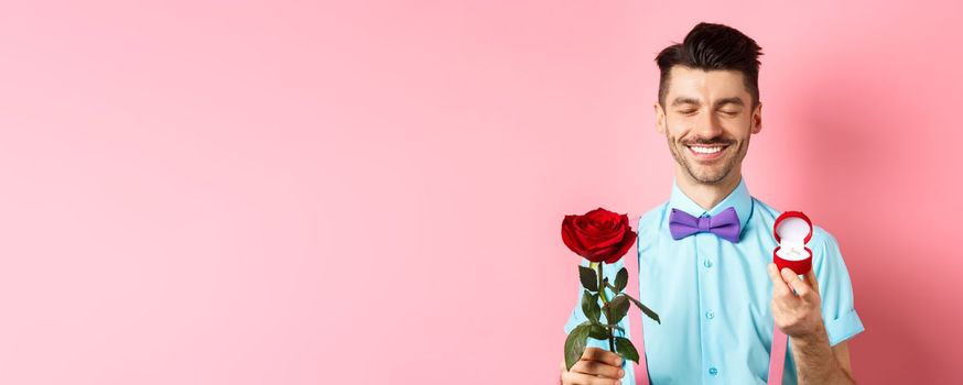 Valentines day. Cute boyfriend making wedding proposal, showing engagement ring in small box and red rose, express love, standing over pink background.
