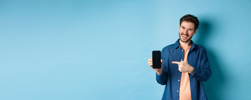 Smiling caucasian man showing on empty smartphone screen, pointing at mobile phone and looking satisfied, standing on blue background.