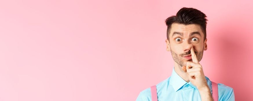 Close-up of funny caucasian man picking nose, staring silly at camera, standing on pink background. Copy space