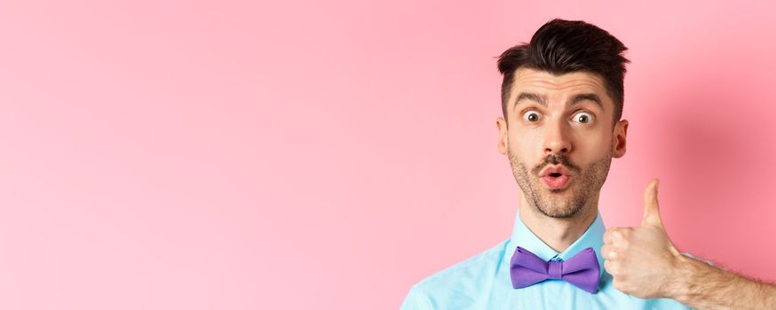 Close-up of funny guy with moustache, say wow and showing thumb up in approval, checking out something cool, recommending product, standing on pink background.