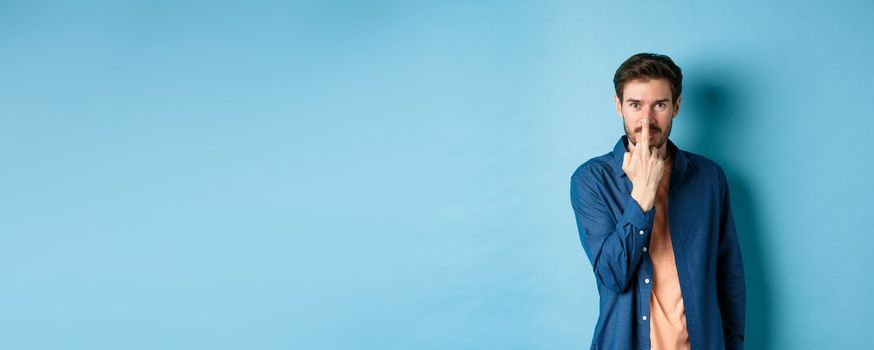 Ignorant and rude guy showing middle finger and smiling, say fuck you, standing on blue background.