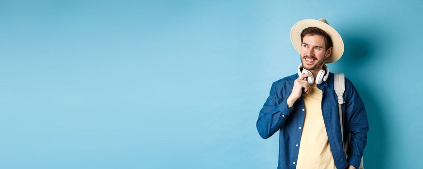 Handsome tourist with backpack and straw hat looking aside, smiling and touching headphones, standing on blue background.