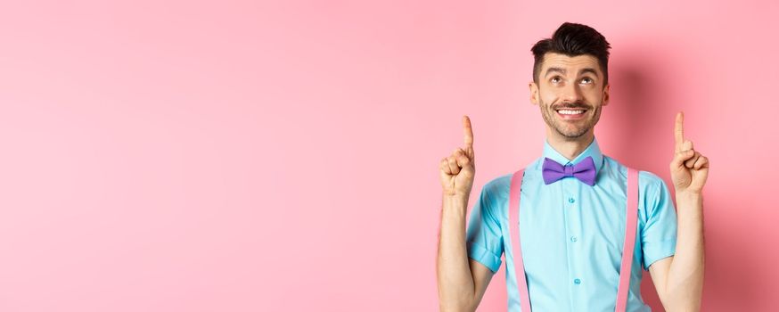 Happy smiling man in bow-tie pointing, looking up, showing something cool, standing on pink background.