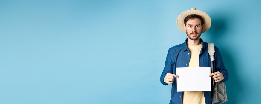 Image of smiling guy in straw hat backpacking, hitchhiking with piece of paper, travelling abroad on summer vacation, blue background.