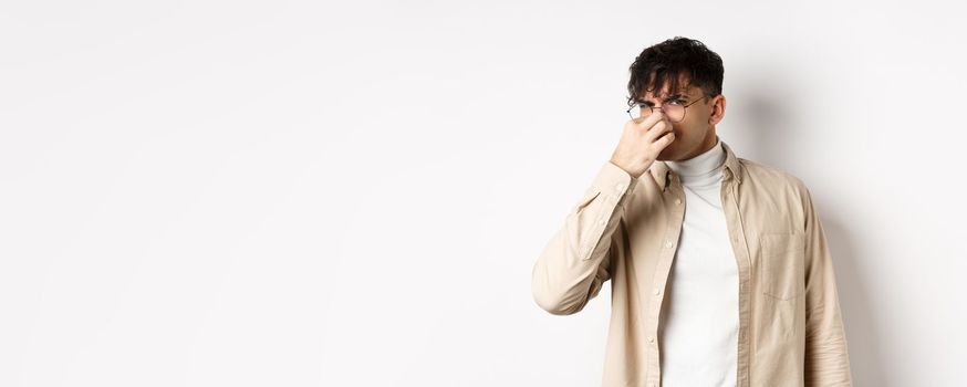 Image of disgusted guy shut his nose from awful smell, looking at something disgusting and stinky, standing on white background.