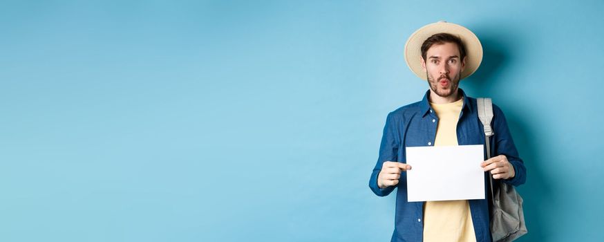 Excited tourist in straw hat, holding empty piece of paper and looking amused, going on summer travel, standing on blue background.