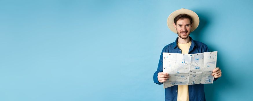 Happy and positive guy on vacation, looking at camera and holding map, smiling excited, going on summer travel, standing on blue background.