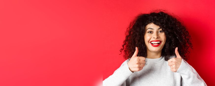 Close up of cheerful caucasian woman showing thumbs up in approval, smiling pleased, like and praise something good, standing on red background.