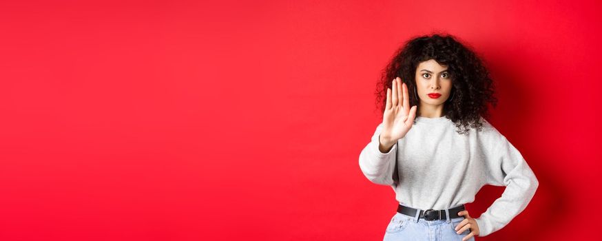 Confident tensed woman extend hand to say stop, disapprove action and prohibit it, make no gesture, standing on red background and forbid something.