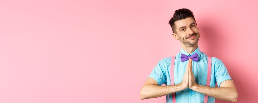 Smiling young guy in bow-tie say thank you, looking with gratitude and holding hands in namaste gesture, standing over pink background. Copy space