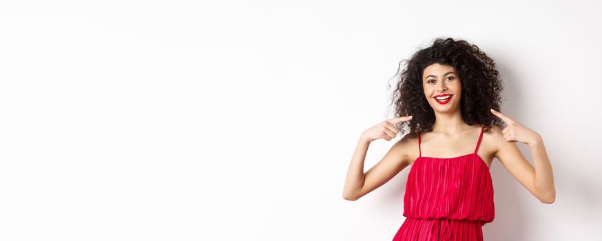 Beautiful woman with curly hairstyle, makeup and red dress, pointing at herself, smiling with white teeth, standing on white background.