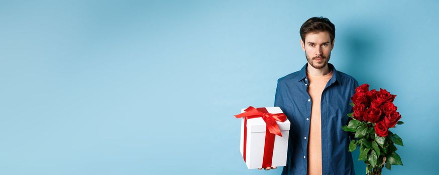 Love and Valentine day concept. Passionate man looking confident at camera, holding gift box and red roses for romantic date, standing over blue background.