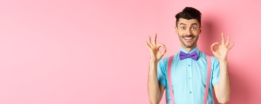 Cheerful handsome man smiling, showing okay gestures in approval, praising good choice, liking advertisement, standing on pink background.