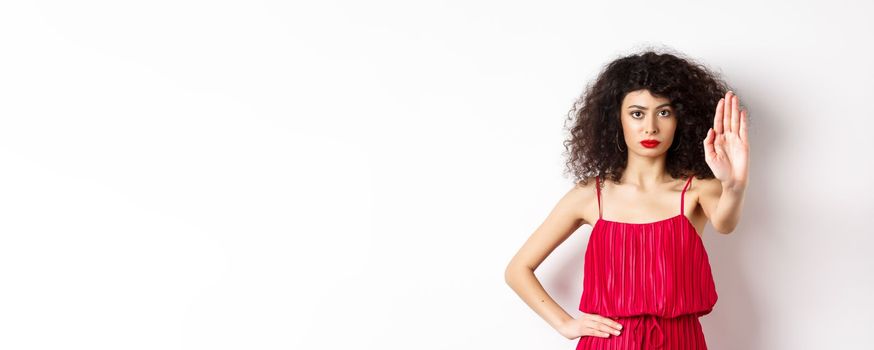 Serious and confident woman in red dress and makeup stretch out hand, tell to stop, prohibit and forbid something, standing over white background.