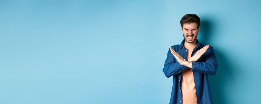 Angry young man frowning and clenching teeth outraged, showing cross gesture to stop or forbid something, standing on blue background.