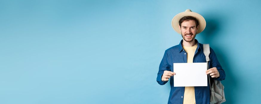 Happy tourist with backpack showing empty piece of paper, smiling at camera, standing on blue background. Concept of summer holiday and vacation.